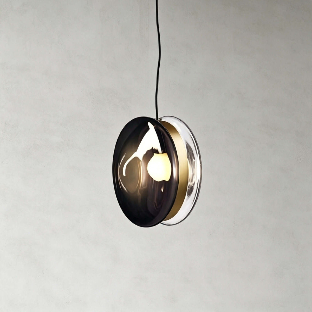This Clérachic glass disc pendant in smoky grey glass, with a brass band and a clear rim, against a grey plaster wall.