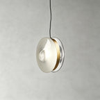 A Clérachic glass disc pendant in milky white glass with a brass band and a clear rim, hanging on a black cord.