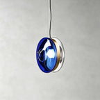 This Clérachic glass disc pendant in deep dark blue glass with a brass band, lit, against a grey plaster wall.
