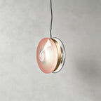 Clérachic glass disc pendant in soft pink glass with a brass band and a clear rim, hanging from a black cord.