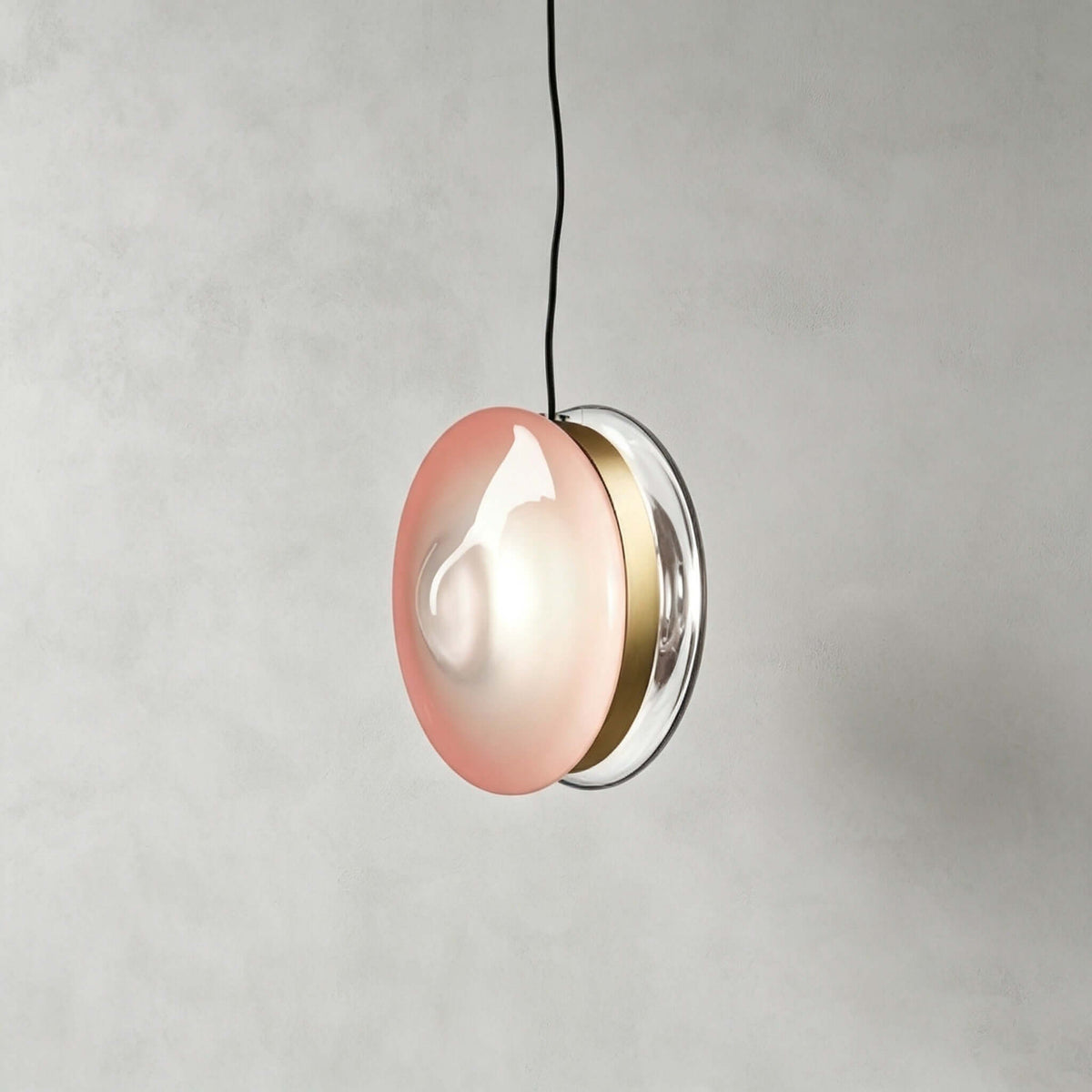 Clérachic glass disc pendant in soft pink glass with a brass band and a clear rim, hanging from a black cord.