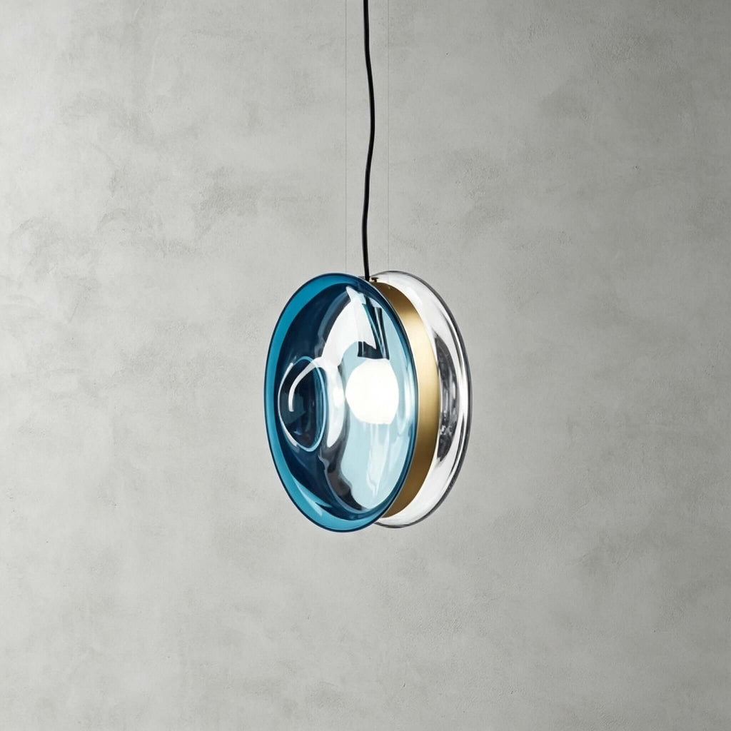 A Clérachic glass disc pendant in light blue glass with a brass band, its bulb glowing, against a grey wall.