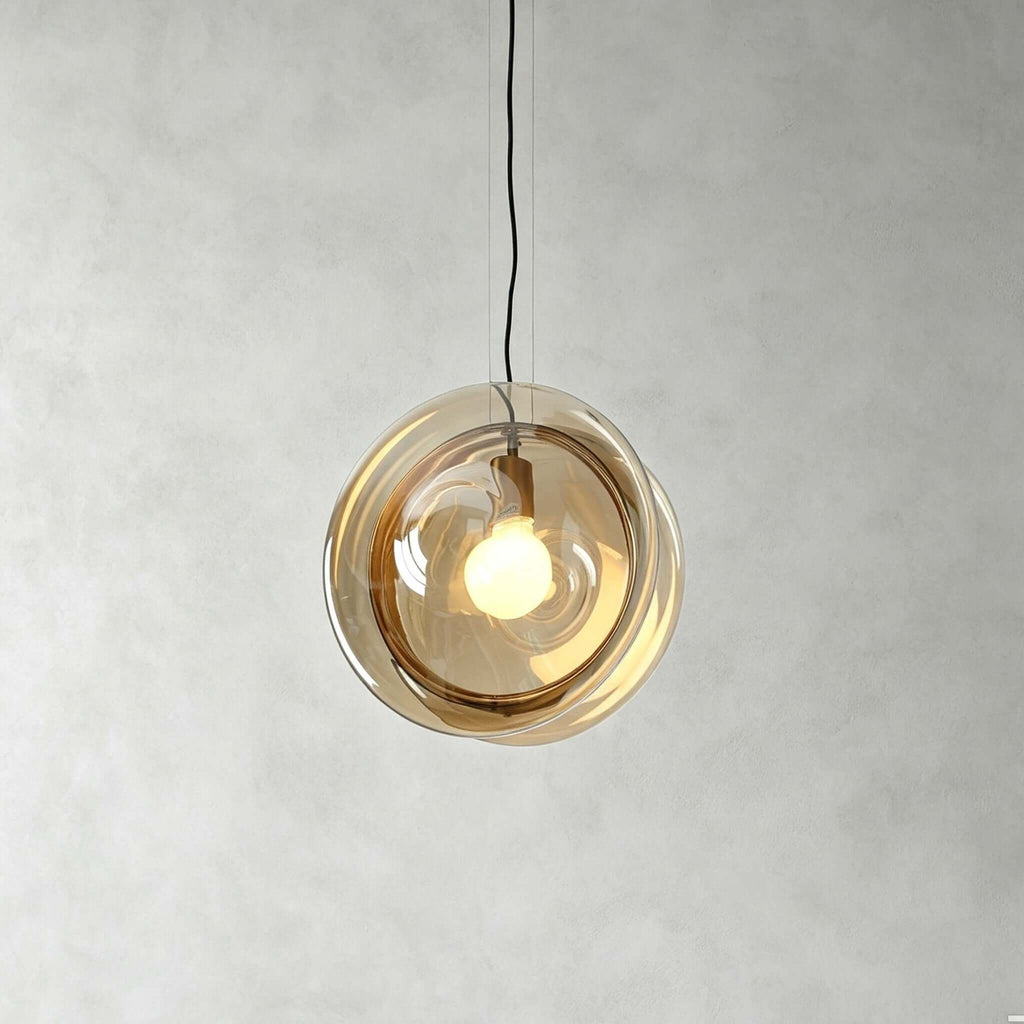 A Clérachic glass pendant in amber glass, lit warmly, hanging from a black cord against a grey plaster wall.