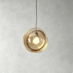 A Clérachic glass pendant in amber glass, lit warmly, hanging from a black cord against a grey plaster wall.