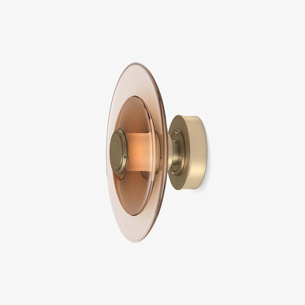 A round Clérachic wall light with a blush-tinted glass disc and a gold back plate, shown at an angle on a plain white backdrop.