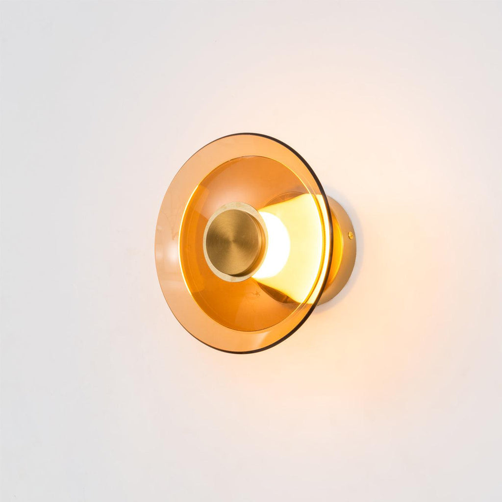 A lit Clérachic wall light with an amber glass disc and a gold centre cap, glowing warm against a pale wall.