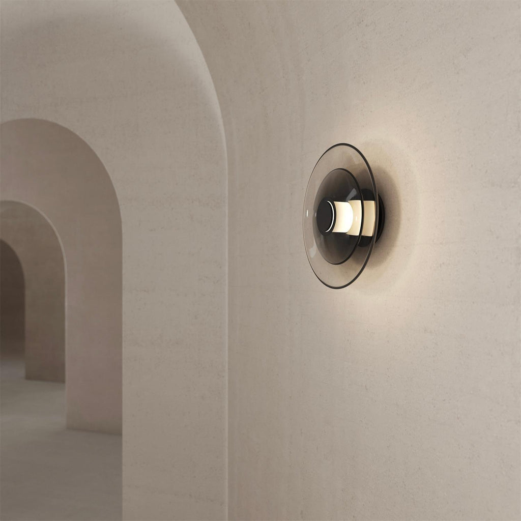 A Clérachic wall light with a smoked-glass disc glows softly on a stone-textured wall beside a row of arched openings.