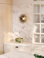 A Clérachic disc wall light with smoked glass hangs on a white marble wall beside a glass-fronted cabinet and a fruit bowl.