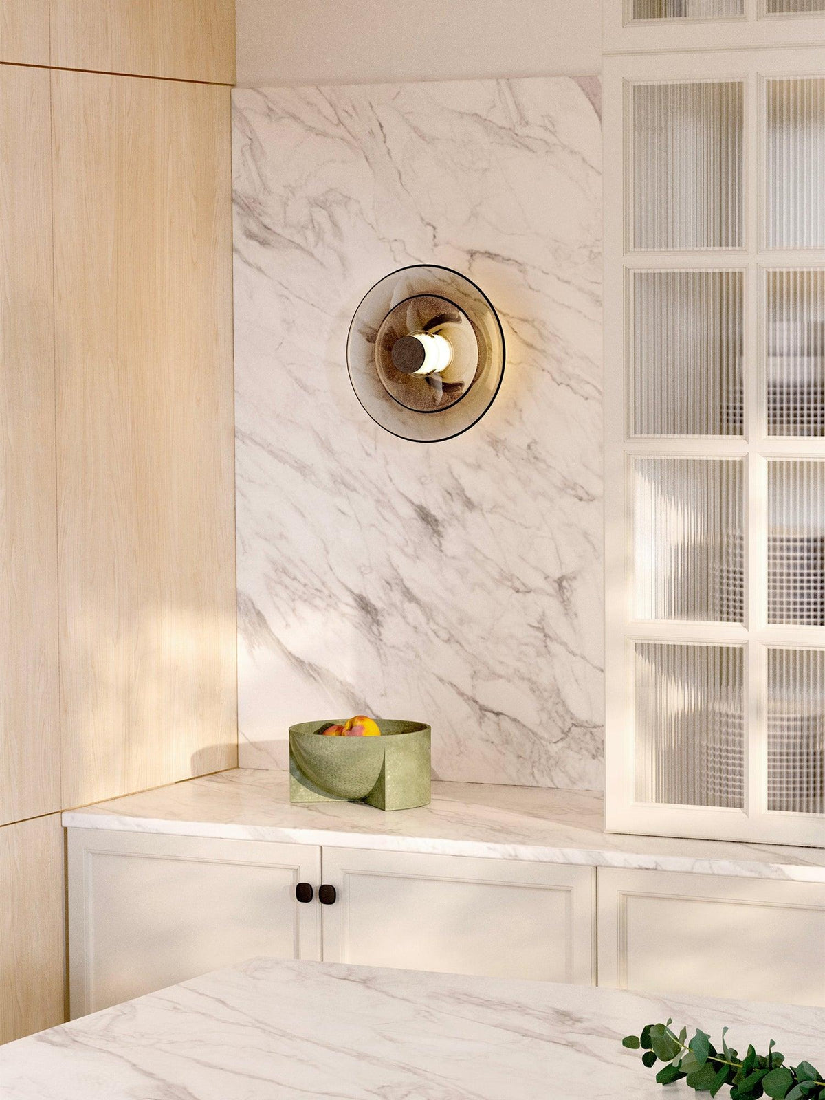 A Clérachic disc wall light with smoked glass hangs on a white marble wall beside a glass-fronted cabinet and a fruit bowl.