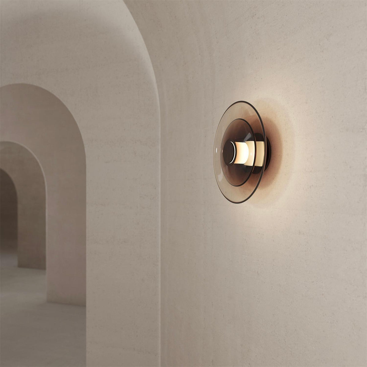 A Clérachic wall light with a dark smoked-glass disc and a lit gold centre on a textured wall, beside tall arches.