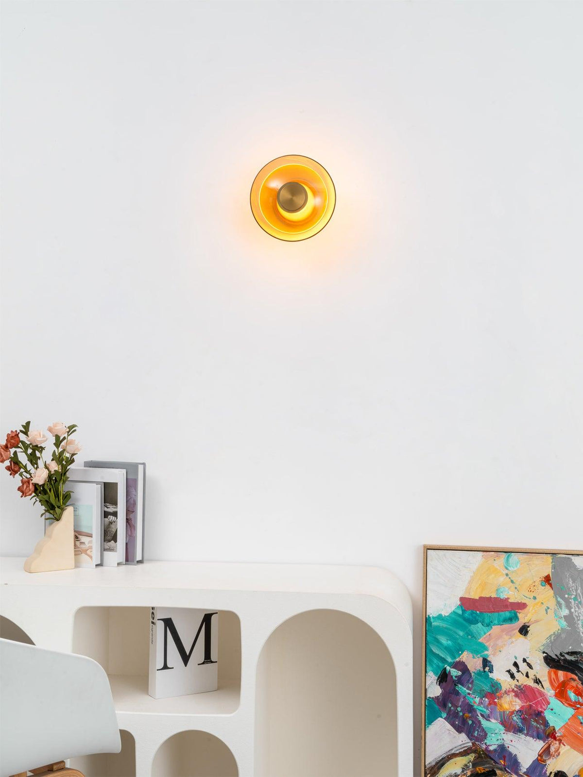 A small Clérachic wall light glows amber on a white wall above a curved white console, a vase of flowers and a colourful print.