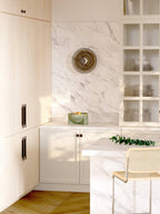 A Clérachic disc wall light on a marble splashback, in a kitchen with pale wood cupboards, a green bowl and a cane stool.
