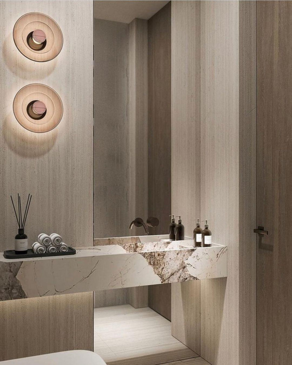 Two Clérachic glass-disc wall lights glow softly above a stone vanity, with reed diffusers and a dark panelled wall.