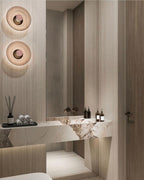 Two Clérachic glass-disc wall lights glow softly above a stone vanity, with reed diffusers and a dark panelled wall.