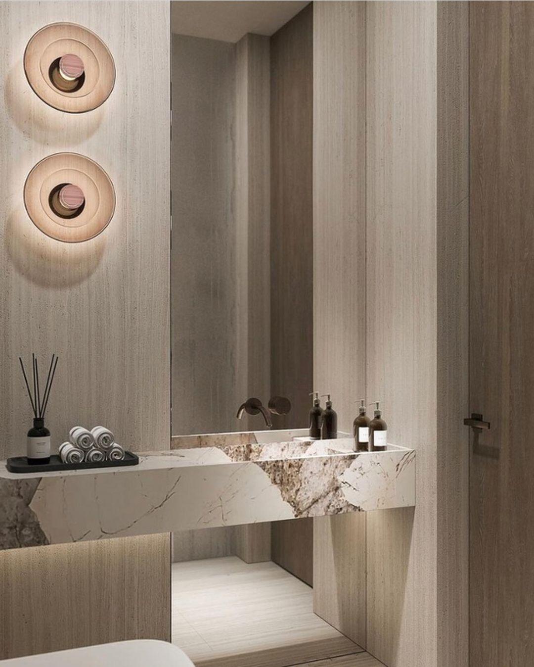 Two Clérachic glass-disc wall lights glow softly above a stone vanity, with reed diffusers and a dark panelled wall.