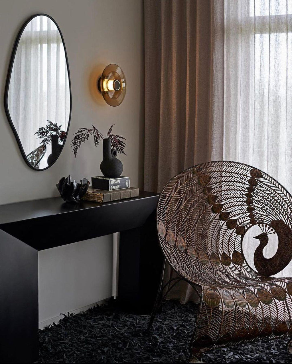 A Clérachic glass-disc wall light glows warm above a black console, beside a mirror and a wire peacock-style chair.