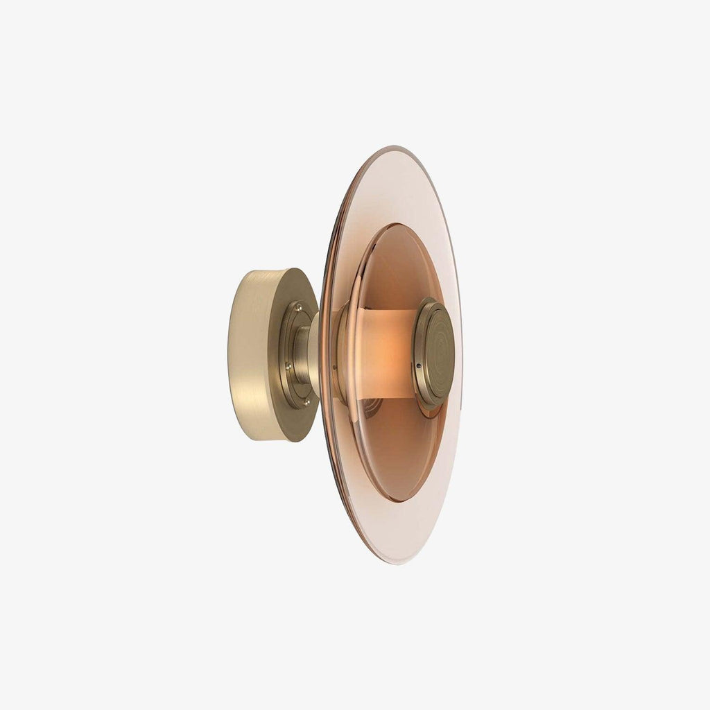 A Clérachic wall light with a pink-tinted glass disc and a gold back plate, seen from the side on a pale backdrop.