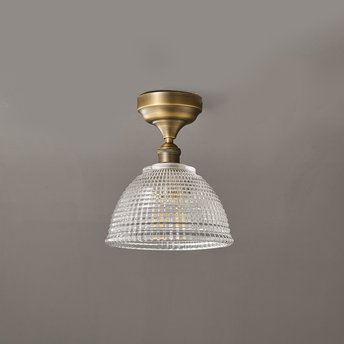 Unlit, the clear grid-patterned glass dome of this Clérachic ceiling light hangs from a short antique brass stem.