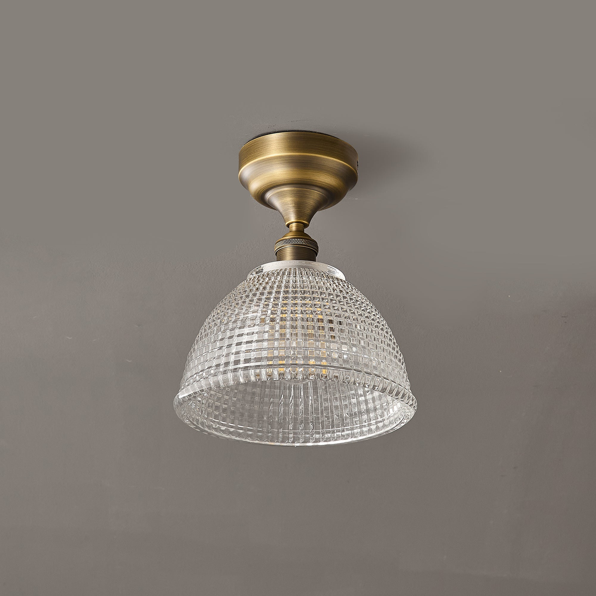 A Clérachic ceiling light seen straight on against a grey ceiling, the clear glass dome hanging below a round brass canopy.