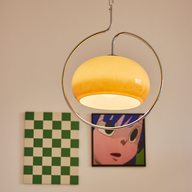 Lit Clérachic glass dome pendant in warm amber, held inside a looping chrome ring, hanging in front of a peach wall with framed prints.