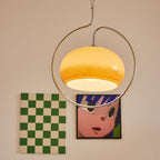 Lit Clérachic glass dome pendant in warm amber, held inside a looping chrome ring, hanging in front of a peach wall with framed prints.