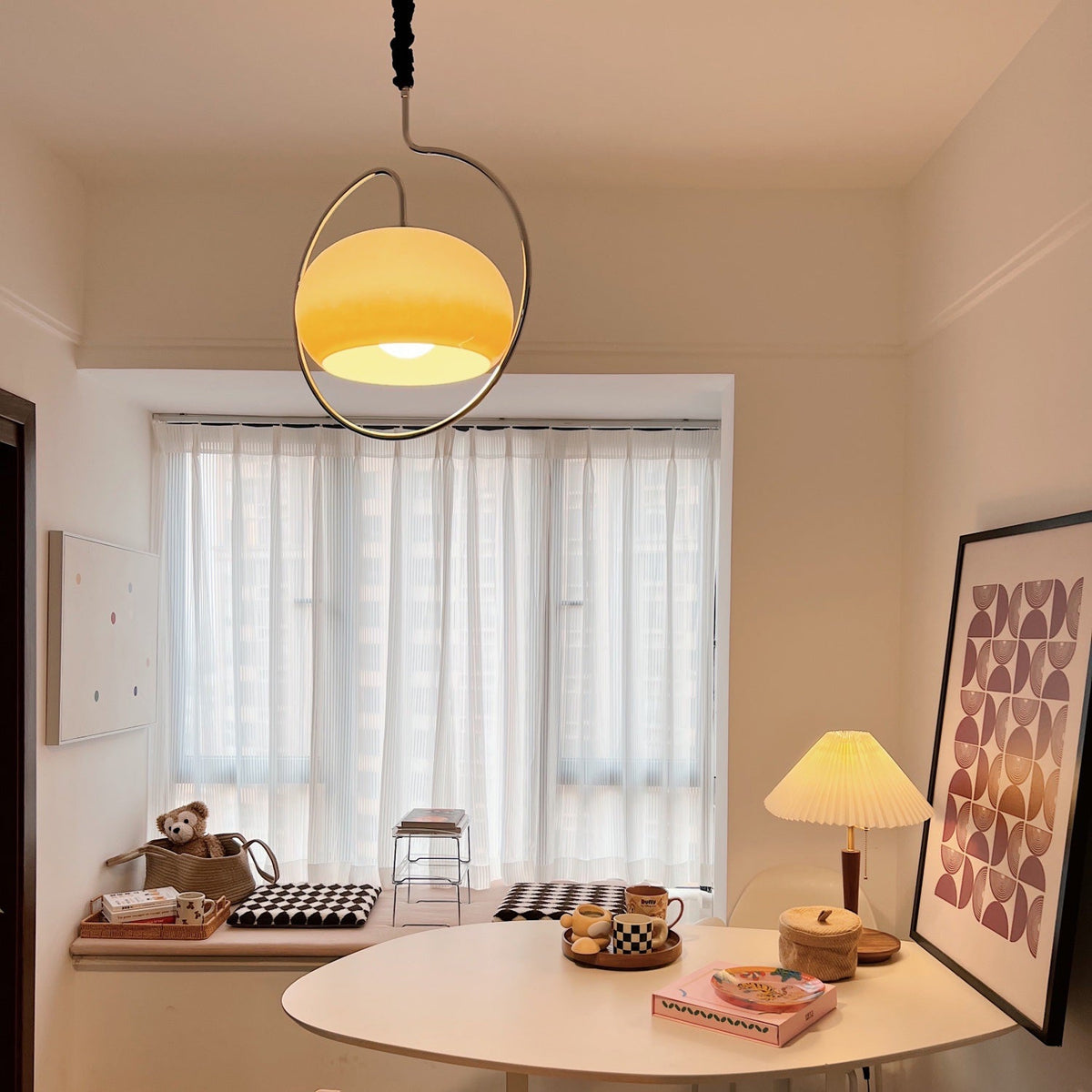 Small Clérachic pendant lit amber inside its chrome ring, above a round white table in a room with sheer curtains and a pleated table lamp.