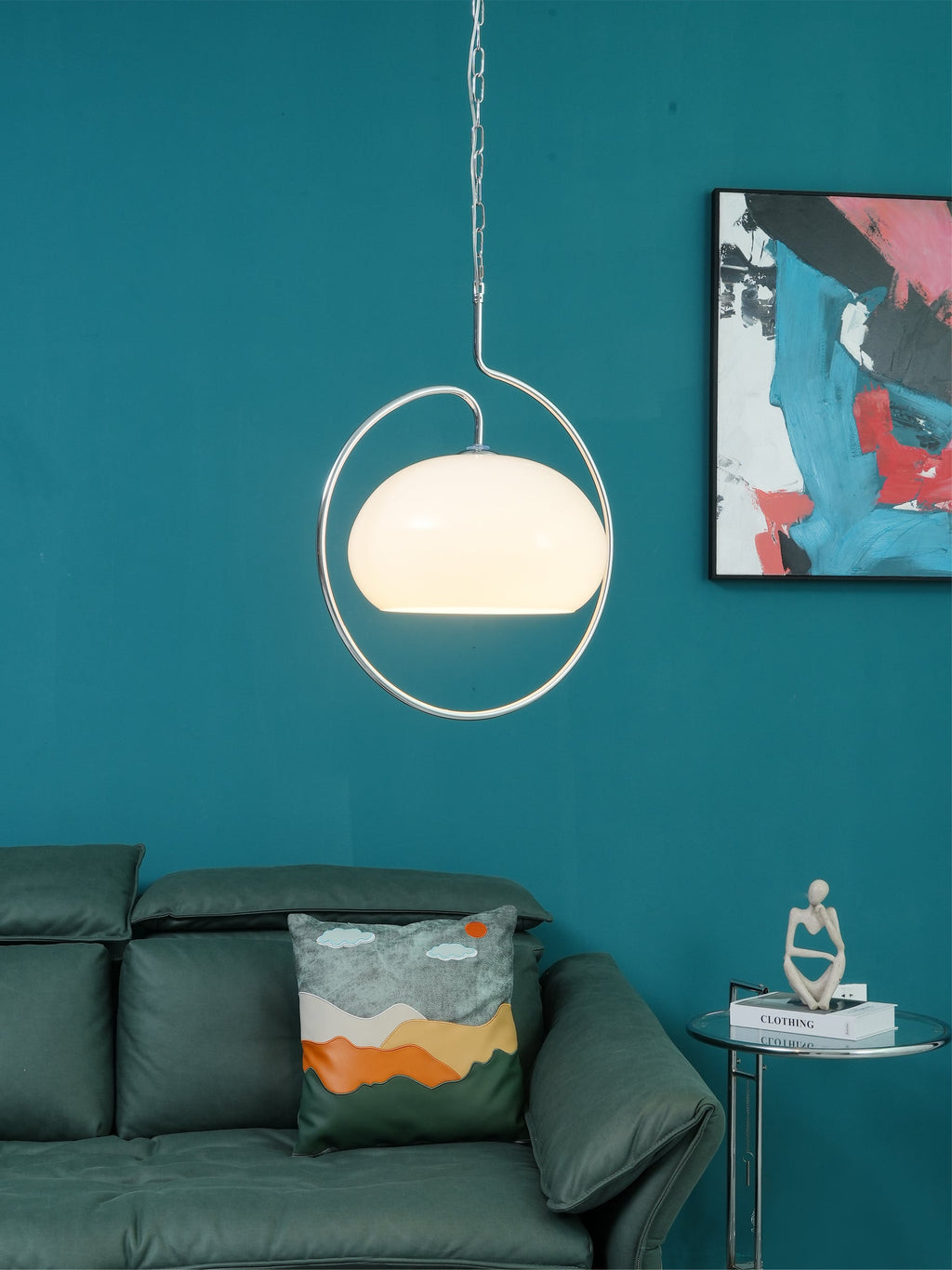 White glass Clérachic pendant lit inside its chrome ring, on a teal wall above a green leather sofa and a glass side table.