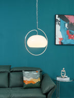 White glass Clérachic pendant lit inside its chrome ring, on a teal wall above a green leather sofa and a glass side table.