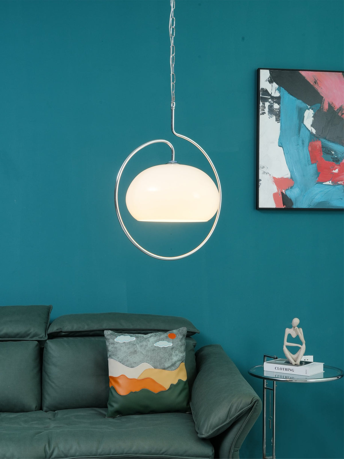 White glass Clérachic pendant lit inside its chrome ring, on a teal wall above a green leather sofa and a glass side table.