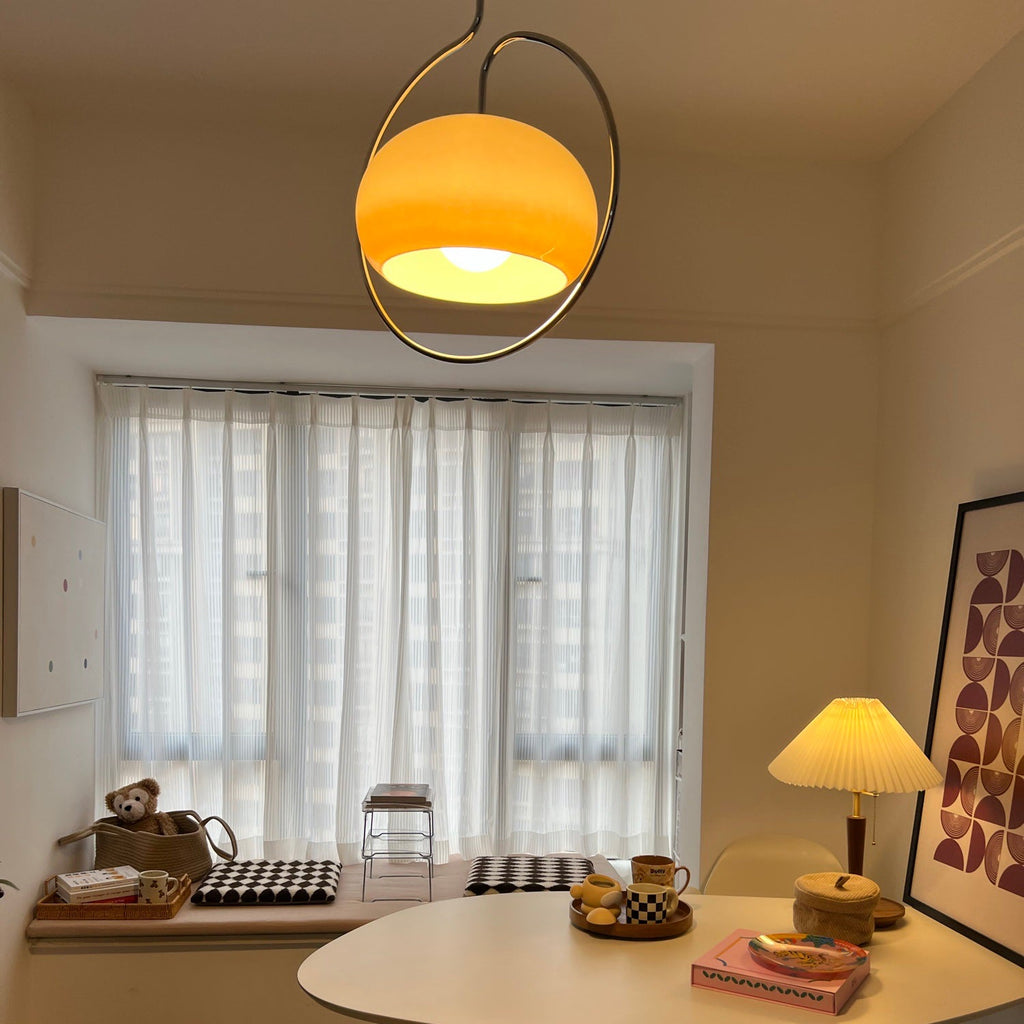 Clérachic pendant glowing orange-amber in its chrome ring, high above a table in a bright room with sheer curtains and a pleated lamp.