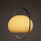 Close view of a lit Clérachic glass dome pendant in warm yellow-cream, encircled by a chrome ring, against a dark grey wall.