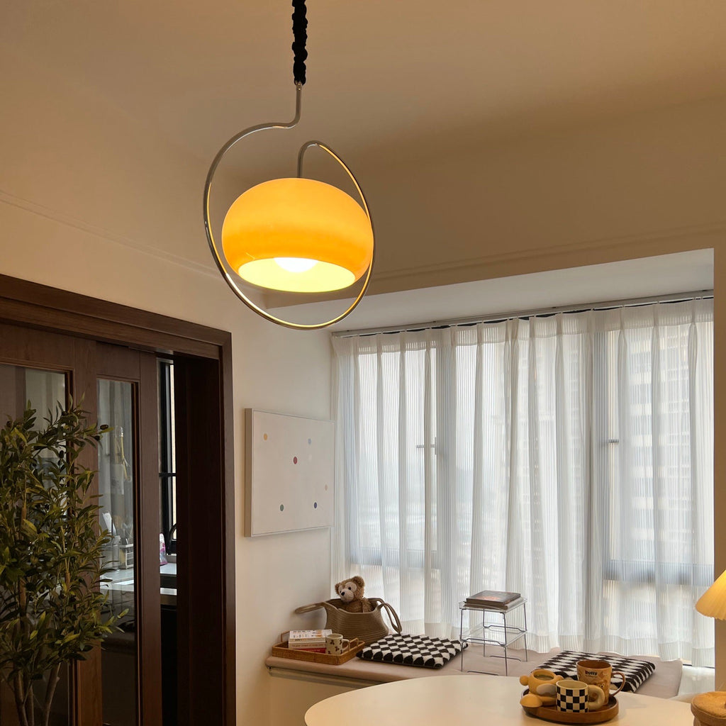 Clérachic pendant glowing amber on a black cord above a dining area with sheer curtains, a dark sideboard and a pleated lamp.