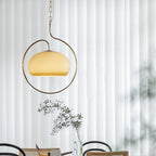 Clérachic pendant with a pale yellow glass dome and a champagne-toned ring, in front of floor-length white curtains and a dining table.