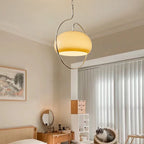 Clérachic glass pendant lit warm white above a bedroom corner, with vertical blinds, a rattan bed and a small framed landscape.