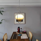Clérachic pendant glowing in front of a framed print on a grey wall, above a wooden dining table with a thermos flask and place mats.
