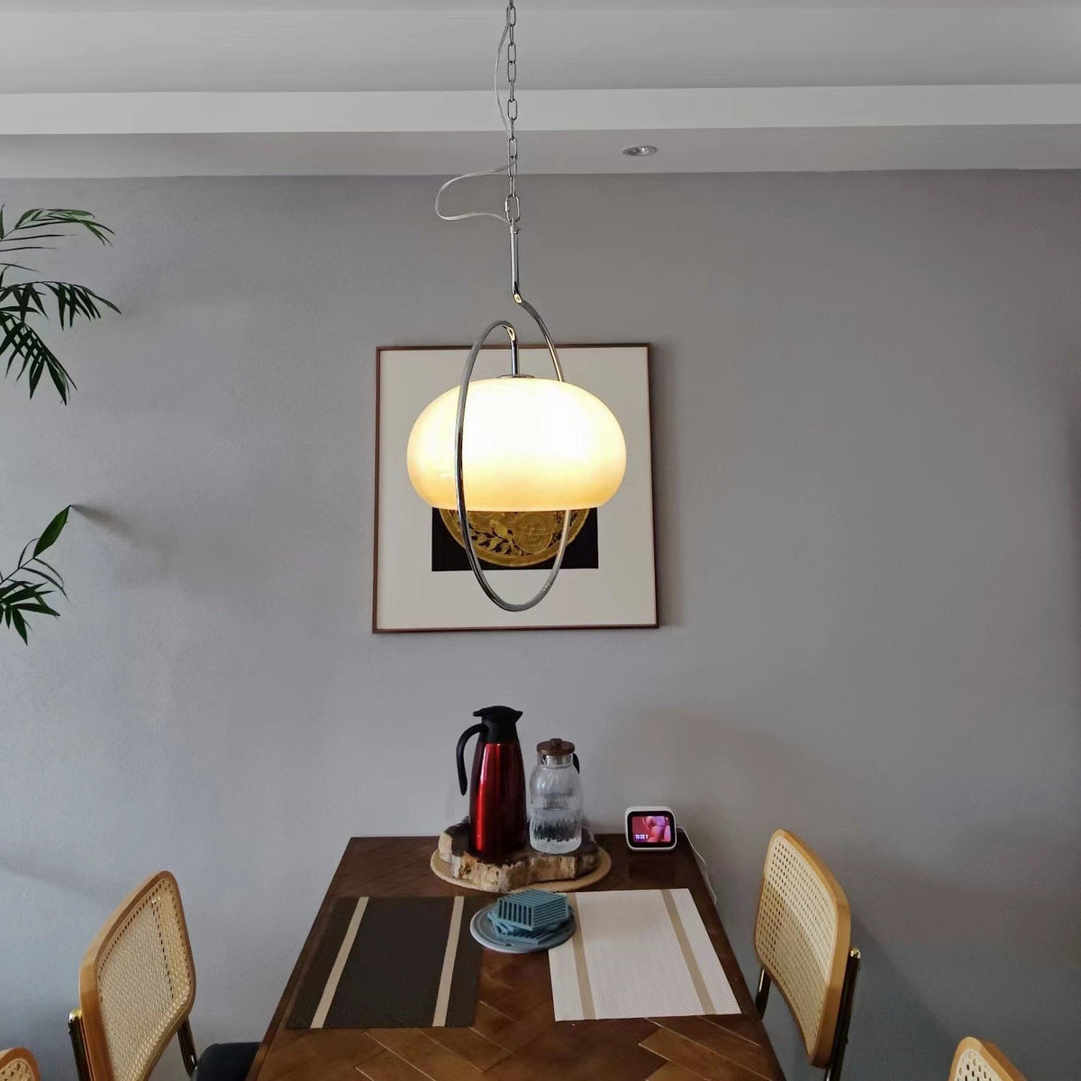 Clérachic pendant glowing in front of a framed print on a grey wall, above a wooden dining table with a thermos flask and place mats.