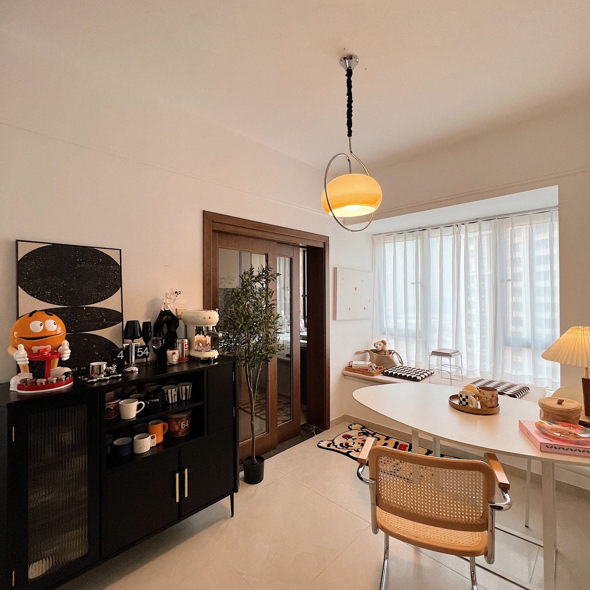 Distant view of a Clérachic pendant glowing amber on a black cord, above a dining room with a dark cabinet and a bay window.