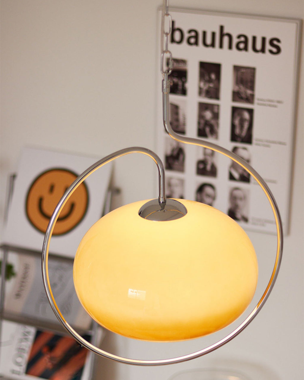 Close view of the lit yellow-cream glass dome and chrome ring of this Clérachic pendant, with a Bauhaus poster and a smiley print behind.