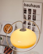 Close view of the lit yellow-cream glass dome and chrome ring of this Clérachic pendant, with a Bauhaus poster and a smiley print behind.