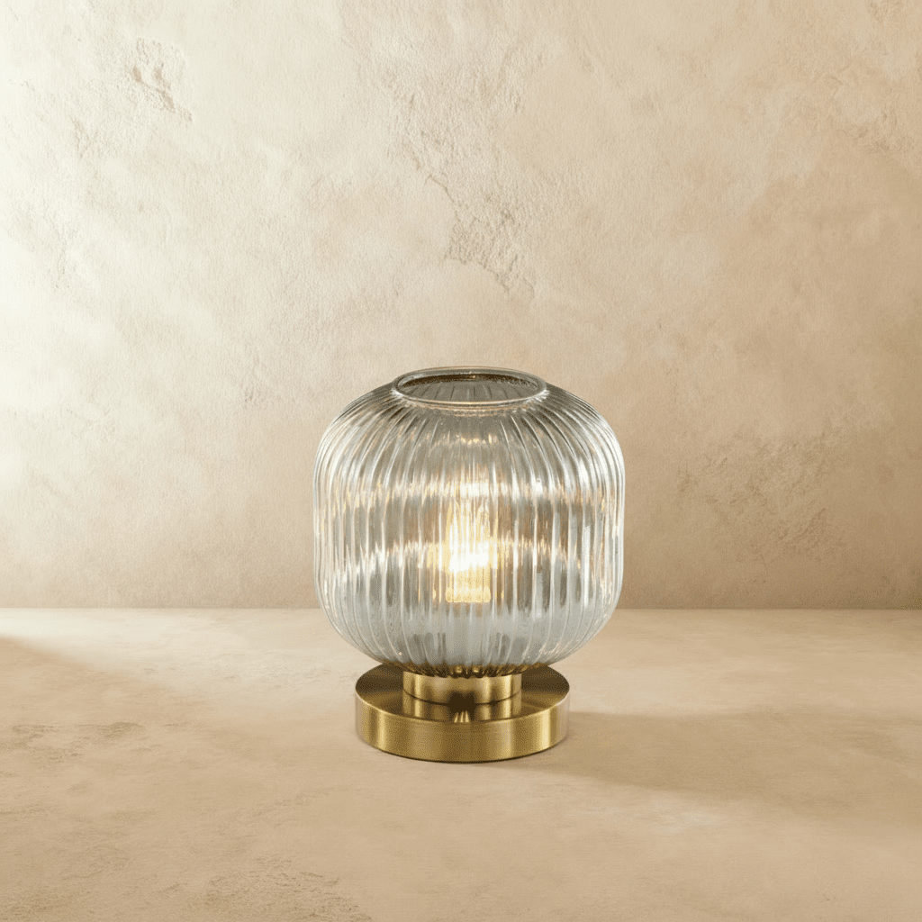 Clérachic ribbed glass dome table lamp in a clear tone, tall silhouette lit warm, brass base on a plaster surface.