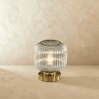 Clérachic ribbed glass dome table lamp in a clear tone, tall silhouette lit warm, brass base on a plaster surface.