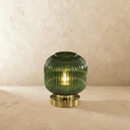 Clérachic ribbed glass dome table lamp in green, small silhouette lit warm, brass base on a plaster surface.