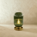 Clérachic ribbed glass dome table lamp in green, tall silhouette lit warm, brass base on a plaster surface.