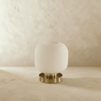 Clérachic ribbed glass dome table lamp in white, small silhouette unlit, brass base on a plaster surface.