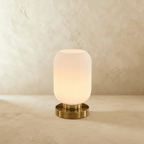 Clérachic ribbed glass dome table lamp in white, tall silhouette lit warm, brass base on a plaster surface.