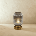 Clérachic ribbed glass dome table lamp in smoky gray, tall silhouette lit warm, brass base on a plaster surface.