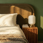 Clérachic ribbed glass dome table lamp in white, tall silhouette lit warm on a fluted-wood nightstand beside a bed.