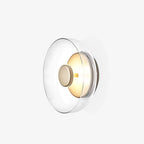 This Clérachic wall light on a plain white backdrop, with a wide clear glass dome over a brass wall plate and a glowing centre disc.