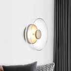 Mounted on a white wall beside a dark grey curtain, this Clérachic wall light has a clear glass dome and a brass centre knob.