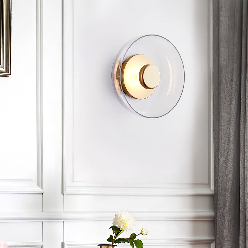 A Clérachic wall light with a clear glass dome and a brass disc hangs on a panelled white wall beside a grey curtain.